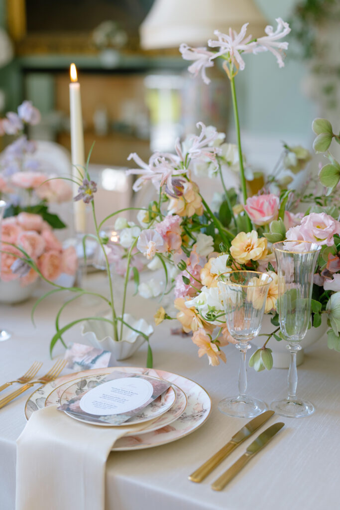 Elegant table setting with floral china, gold cutlery, two glasses, a white menu card, pastel flower arrangements, and a lit candle on a light tablecloth. Soft, natural lighting enhances the romantic atmosphere.