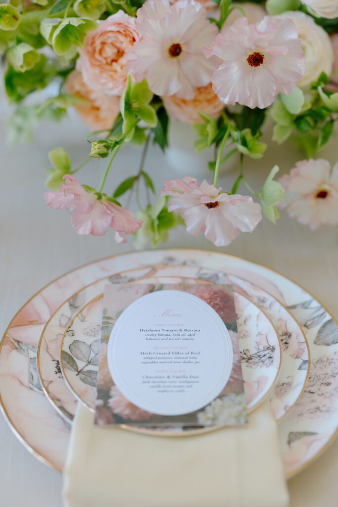 Elegant table setting with floral-patterned plates, a cream napkin, and a printed menu. In the background, soft pink flowers and greenery create a romantic, delicate centerpiece.