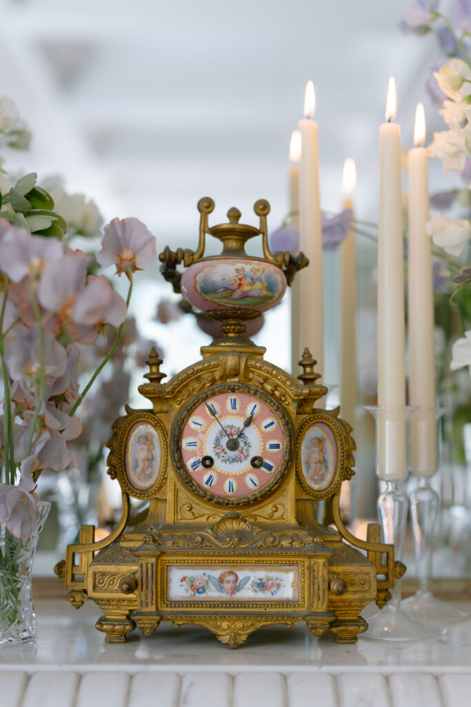 Ornate antique clock with Roman numerals, gold detailing, and painted portraits, surrounded by pastel flowers and tall lit candles on a reflective surface.