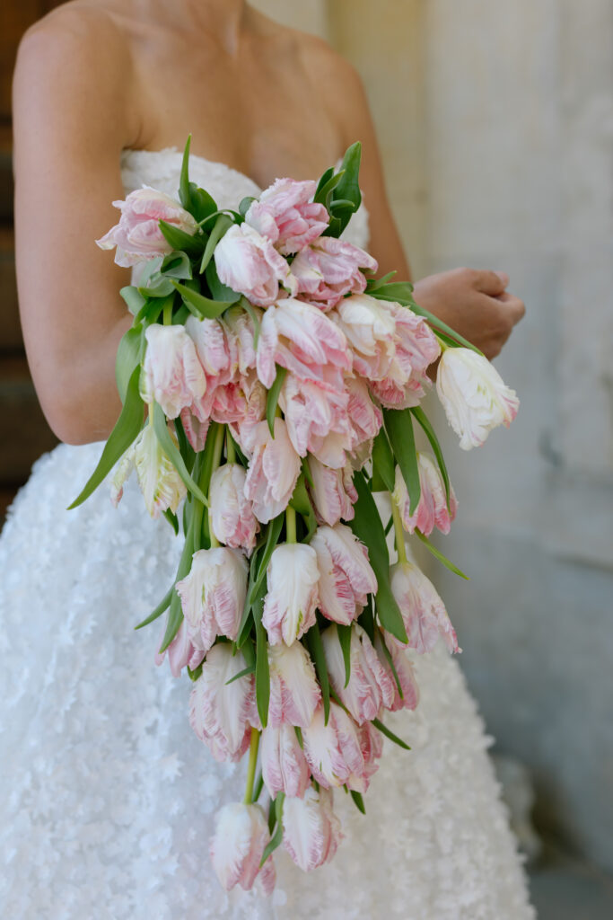 A bride in a white strapless dress holds a large cascading bouquet of pink and white tulips.