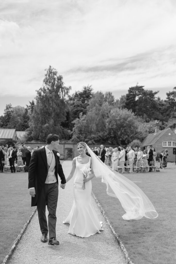A bride and groom walk hand in hand outdoors, smiling. The bride wears a long, flowing veil and holds a bouquet. Guests stand in the background, and trees and buildings are visible behind them. Black and white photo.