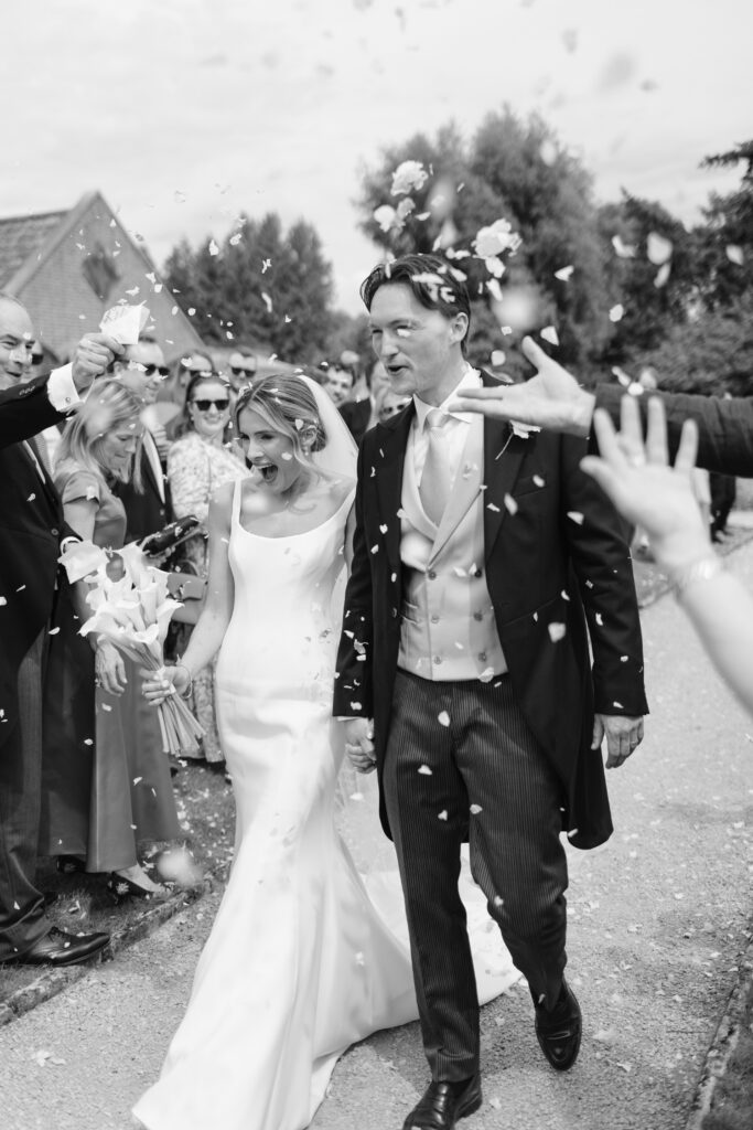 A bride and groom walk hand-in-hand outside as guests throw flower petals. The bride wears a long white dress and veil; the groom wears a suit with a vest and tails. Everyone smiles, celebrating the newlyweds.