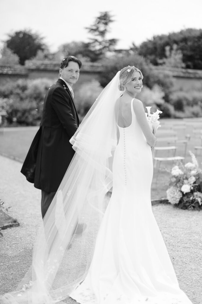 A bride in a long white dress and veil smiles at the camera while holding flowers, standing next to a groom in a suit outdoors. They appear to be walking in a garden setting.