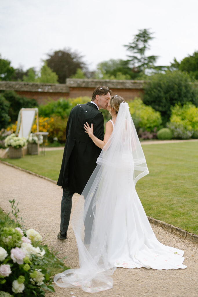 A bride and groom walk hand in hand along a garden path, sharing a kiss. The bride wears a white dress and veil, while the groom wears a black suit. Lush greenery and flowers surround them.