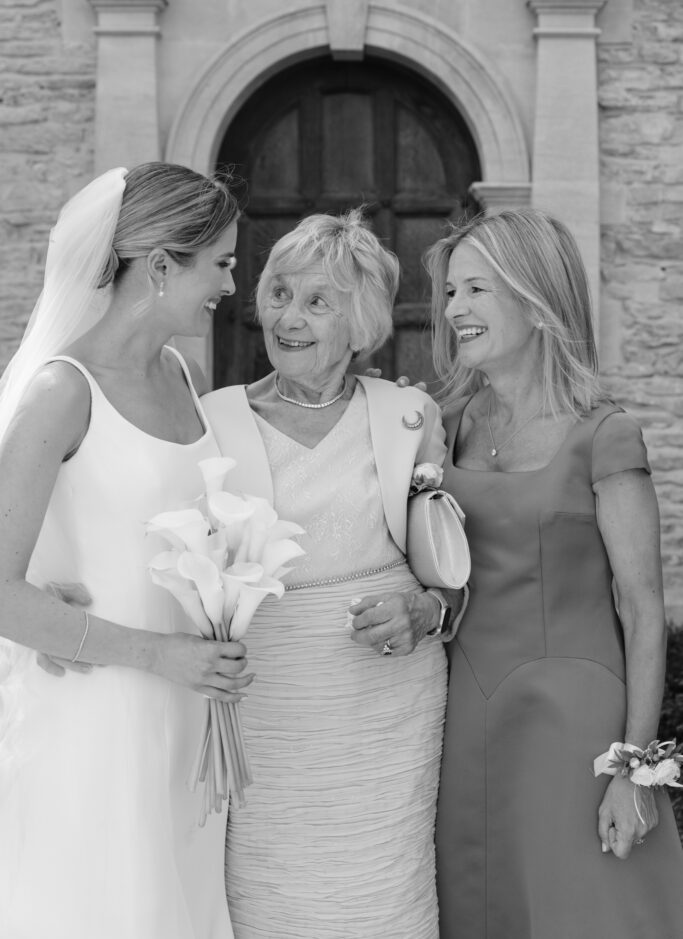 Three women dressed in formal attire stand outside a stone building, smiling at each other. One wears a wedding dress and holds flowers; the others, likely family, wear elegant dresses.