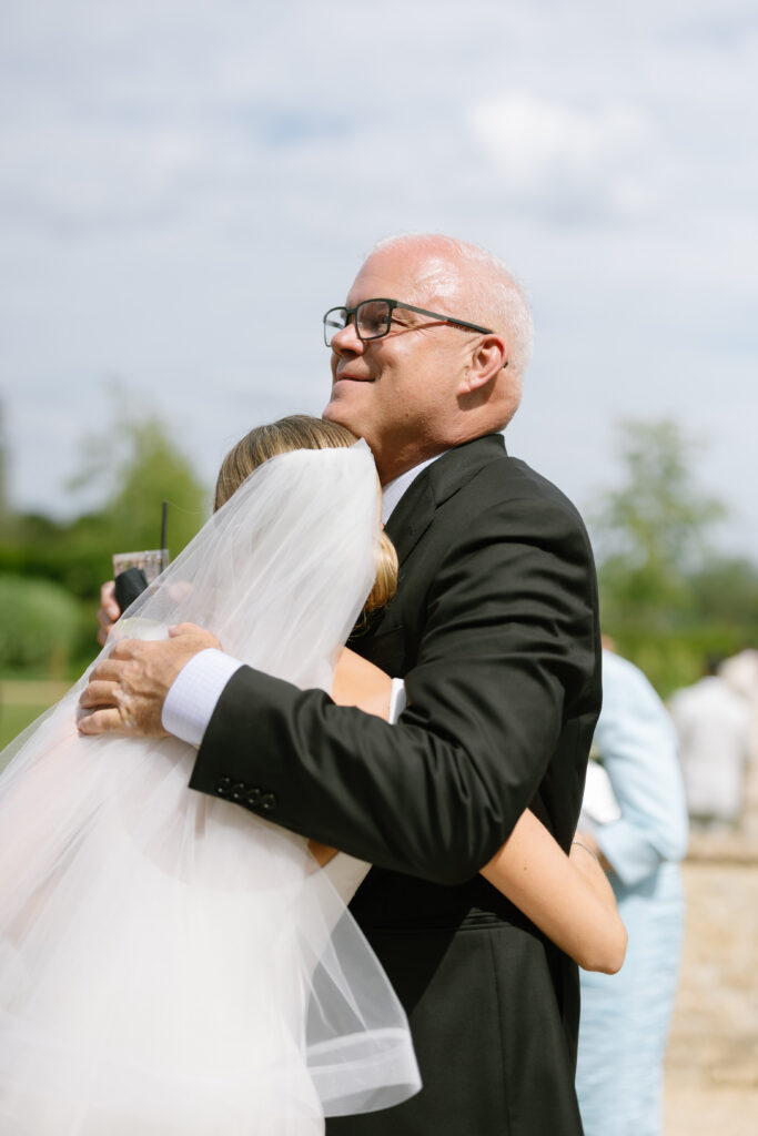 A man in a suit warmly hugs a bride in a white dress and veil outdoors, with a soft background of greenery and a few blurred people.