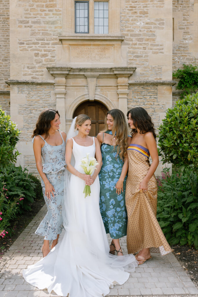 Four women stand outside a stone building. The bride, in a white gown holding flowers, stands in the middle, flanked by three women in colorful dresses. They all smile at each other in a garden setting.