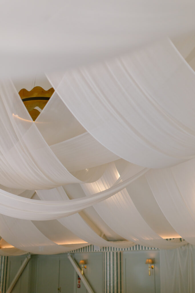 White sheer fabric drapes gracefully from the ceiling, creating a soft, elegant look in a light-filled room with wall sconces and blue-green walls—an exquisite setting reminiscent of a Kin House Wedding.