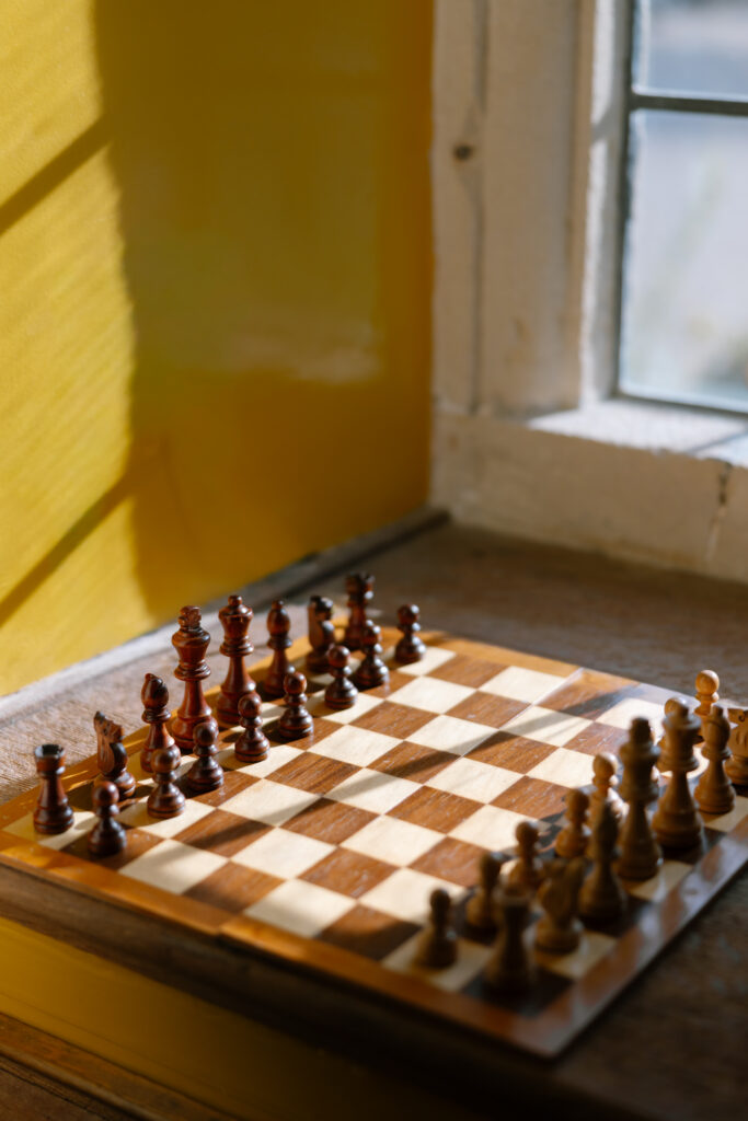 A chessboard with pieces arranged for a new game sits on a wooden windowsill, sunlight streaming in through a nearby window and casting shadows on the yellow wall.