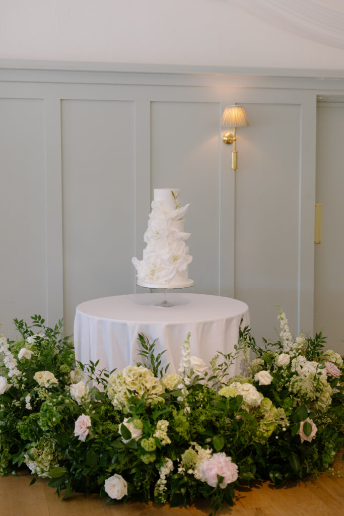 A tall white wedding cake with floral decorations sits on a round table covered with a white cloth, surrounded by lush white and green flower arrangements. The background features pale gray paneling and a wall sconce.