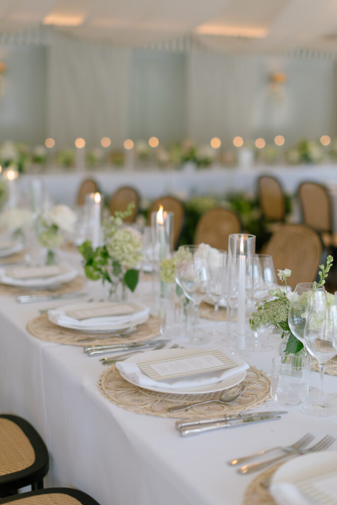 Elegant table setting with white plates, menus, glassware, floral centerpieces, and candles on a white tablecloth, with woven placemats and wooden chairs, beautifully arranged for a formal event or Kin House Wedding reception.