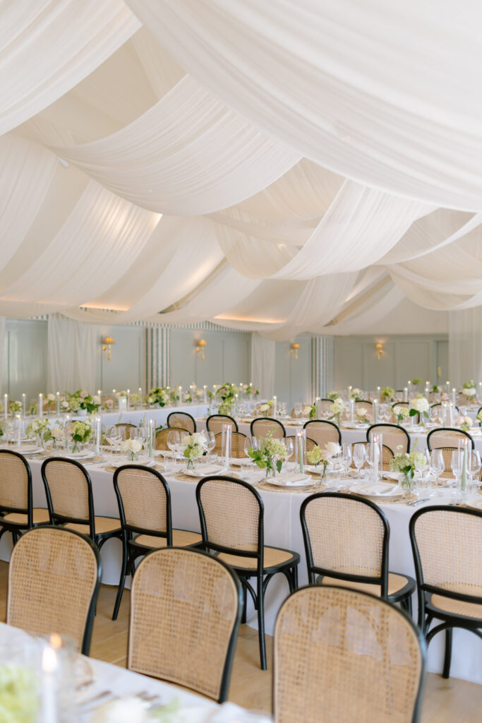 A bright, elegant event hall with long tables set for dining, decorated with green and white flowers, candles, and draped white fabric ceiling. Cane-backed chairs are neatly arranged around the tables.