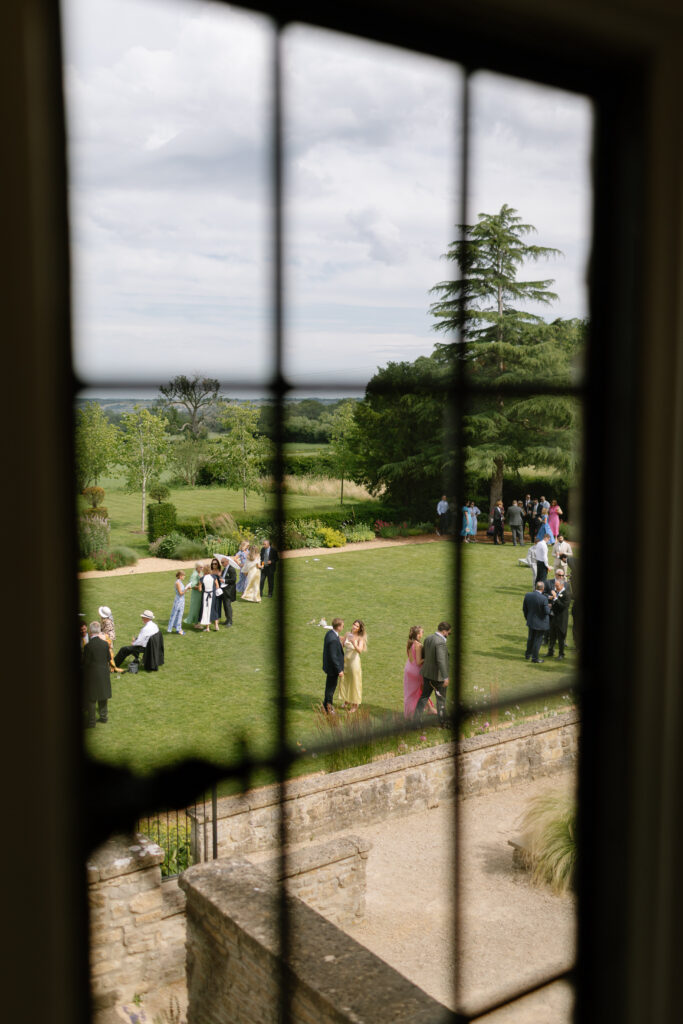 View through a window of people in formal attire socializing on a green lawn with trees and countryside in the background, under a cloudy sky.