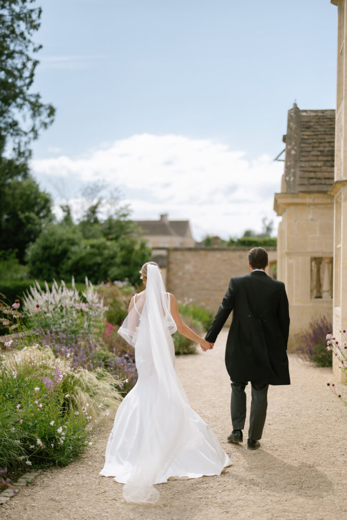 A bride and groom walk hand in hand along a garden path, the bride in a white dress and veil, and the groom in a dark suit, with greenery and stone buildings in the background.