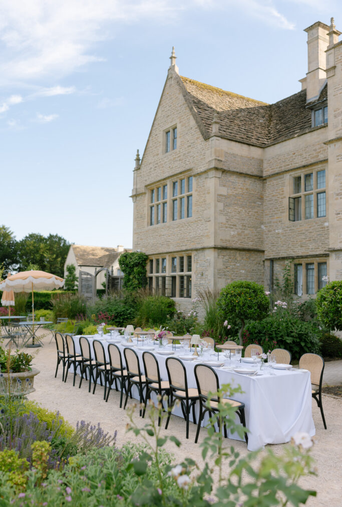 A long, white-clothed dining table with black chairs is set outdoors beside a stone manor house, surrounded by greenery and flowers under a clear blue sky.