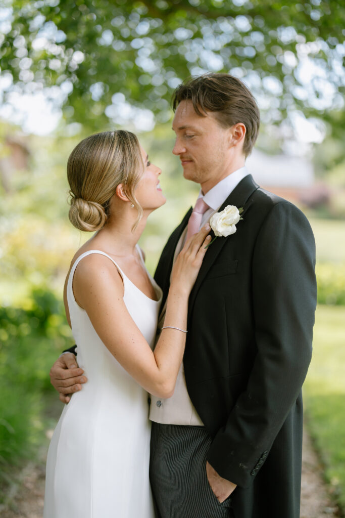 A bride in a white dress and a groom in a suit stand outdoors, embracing and gazing at each other lovingly under green trees.