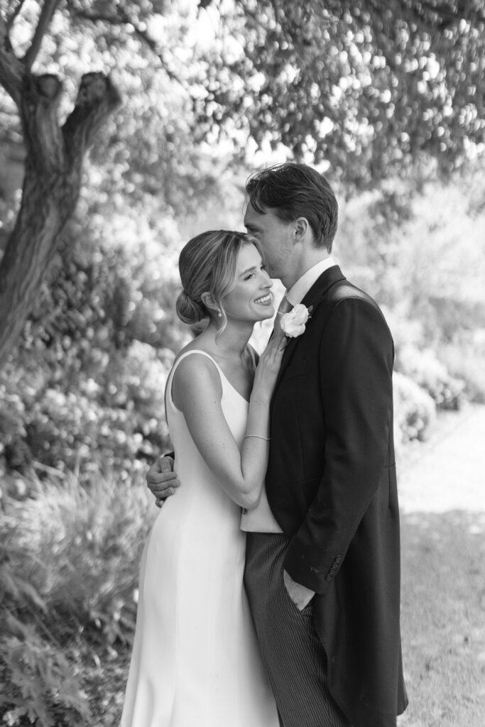 A bride and groom embrace and smile in a garden. The bride wears a sleeveless dress and the groom wears a tuxedo with a boutonnière. The photo is in black and white.
