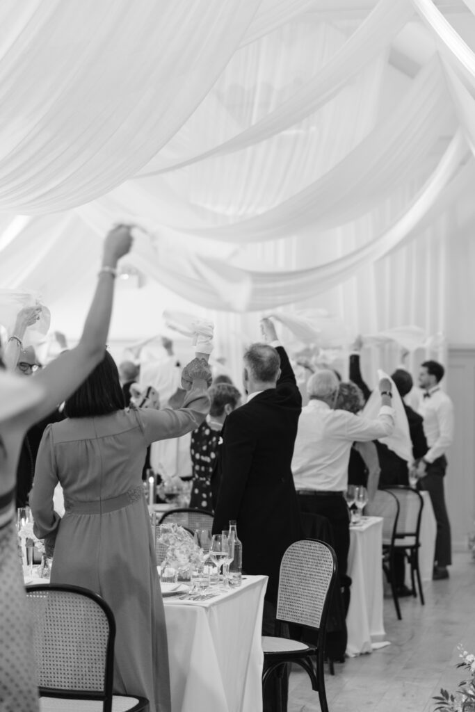 Guests at a formal event stand and wave napkins in the air, celebrating together in a decorated venue with draped white fabric overhead.