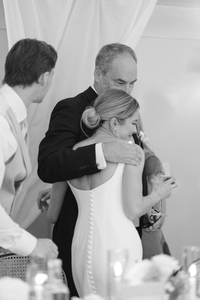 A man hugs a woman in a wedding dress while she holds a wine glass. Another man in a vest stands nearby. The scene is warm and celebratory, with soft lighting and draped fabric in the background.