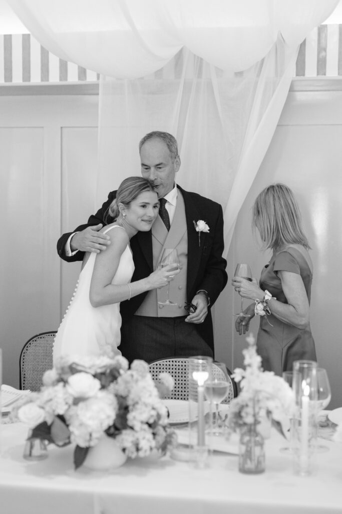 A bride hugs an older man in a suit while a woman in a dress stands nearby. All hold drinks. The setting is elegant with floral centerpieces and sheer white drapery hanging in the background.