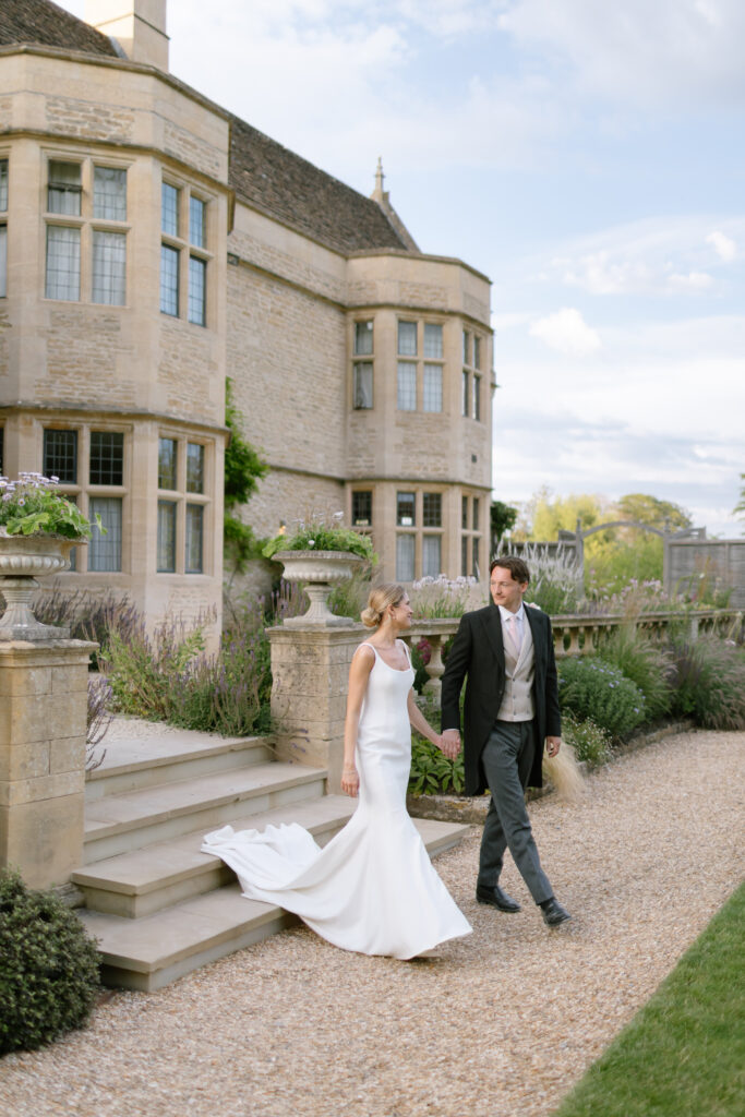 A bride in a white dress and a groom in formal attire walk hand in hand outside a large stone building with tall windows and manicured gardens.
