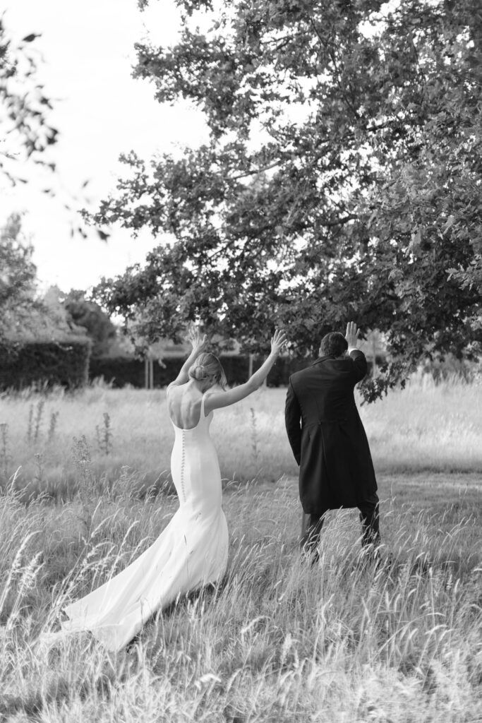 A bride and groom, seen from behind, walk through tall grass with their arms raised, under a large tree. The scene is in black and white.