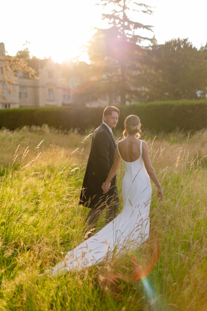 A bride and groom walk hand in hand through a sunlit field, the bride in a white dress with a long train and the groom in a dark suit, with trees and a building in the background.