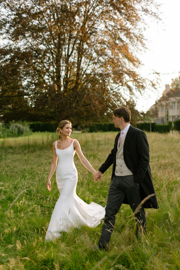 A bride in a white dress and a groom in a suit walk hand in hand through tall grass, smiling at each other, with trees and a house in the background on a sunny day.
