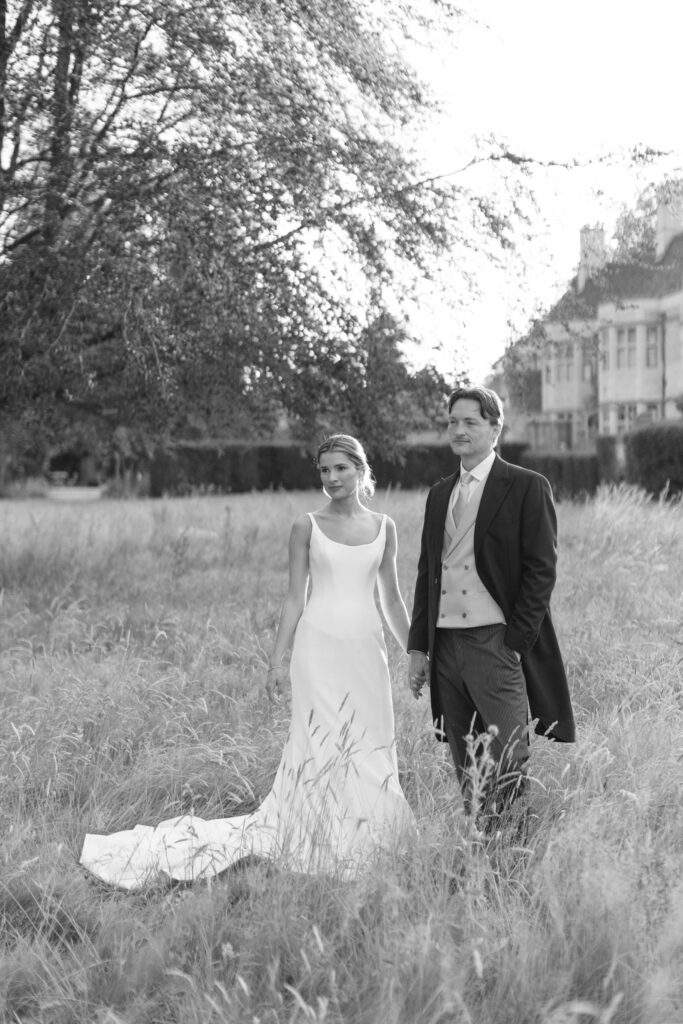 A bride in a long white dress and a groom in formal attire stand in tall grass outdoors, with trees and a large building in the background. The image is black and white.