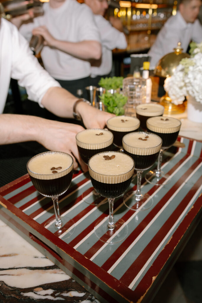 Eight espresso martinis garnished with coffee beans are arranged on a striped tray at a bar, with bartenders in white shirts preparing drinks in the background.