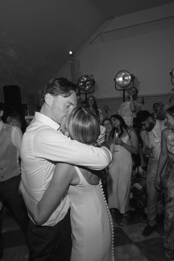 A couple embraces and kisses on a dance floor, surrounded by smiling guests in formal attire. The scene is joyful, with people celebrating and laughing in a lively, softly lit room.