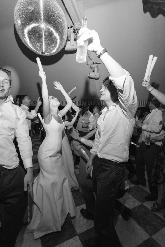 A group of people dance energetically at a party. A woman in a white dress and others raise their arms, some holding light sticks or drinks, under a sparkling light fixture. The scene is lively and joyful.