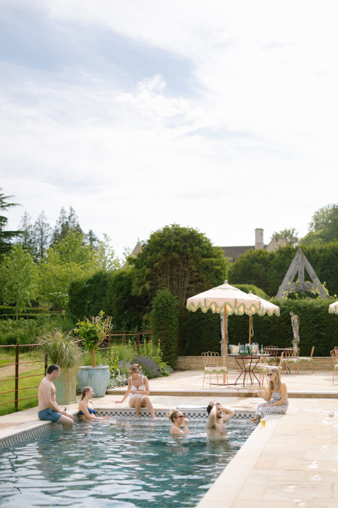 People relax and chat in an outdoor swimming pool beside lounge chairs and umbrellas, surrounded by greenery and a garden on a sunny day.