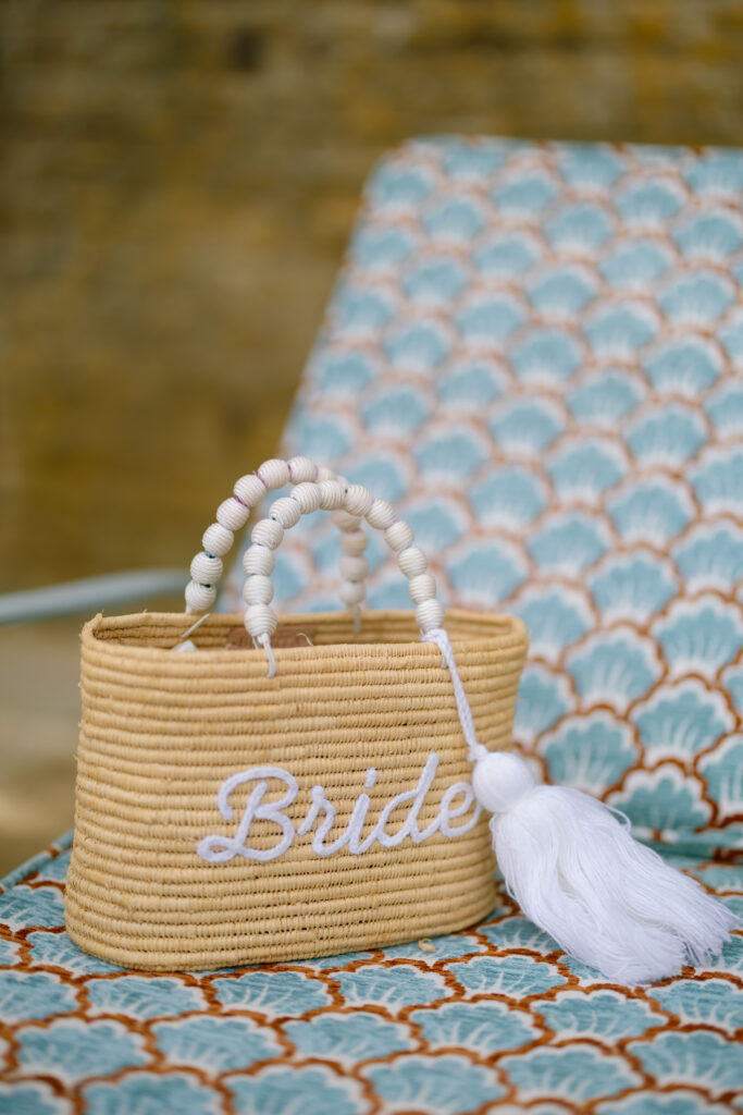 A woven straw bag with white beaded handles, a large white tassel, and the word Bride embroidered on the front sits on a patterned blue and orange chair.