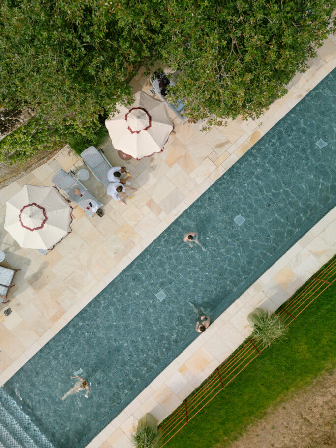 Aerial view of a narrow swimming pool with three people swimming, poolside lounge chairs, umbrellas, and a few people sitting under the umbrellas near trees and greenery.