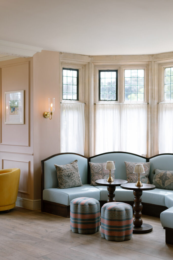 A cozy sitting area with light blue sofas, patterned pillows, round wooden tables, two striped ottomans, and large windows with sheer curtains letting in natural light.