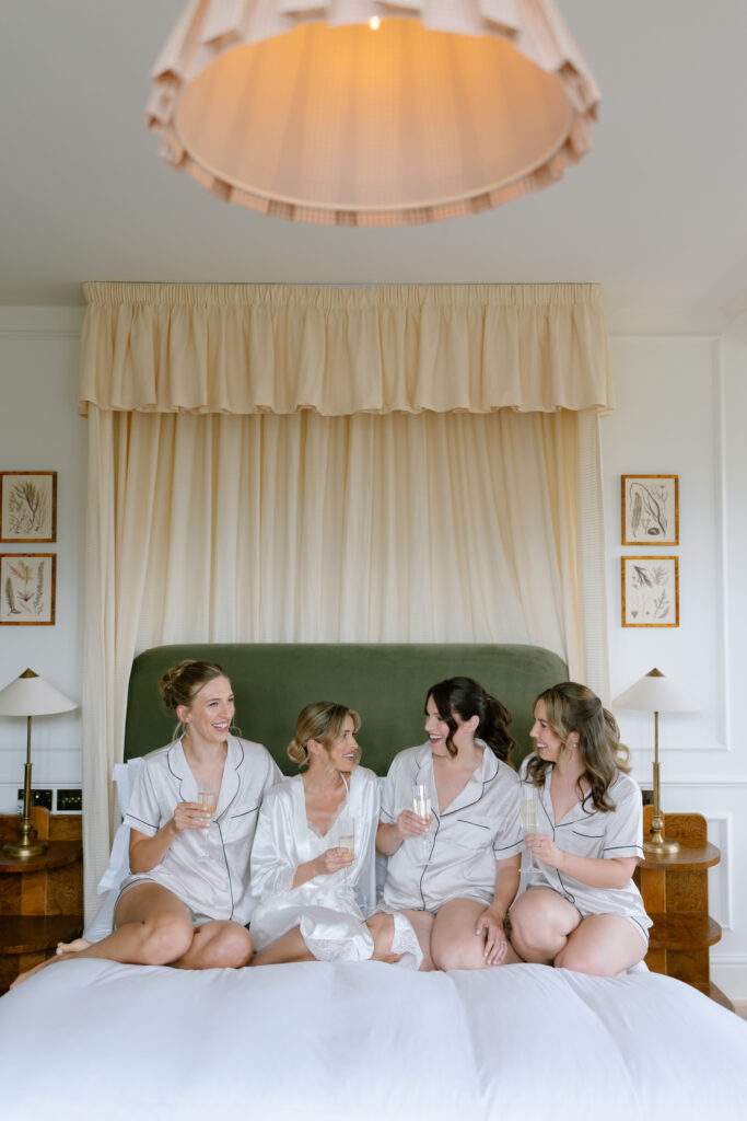 Four women sit on a bed in matching pajamas, smiling and holding glasses of champagne, with a large light fixture hanging above and elegant decor in the background.