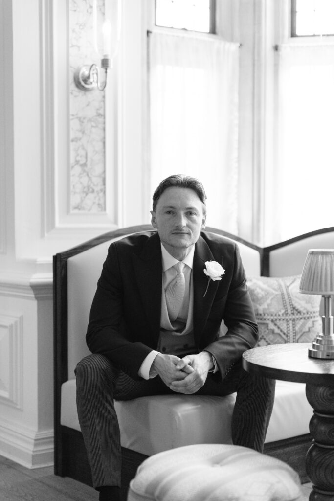 A man in a suit with a boutonnière sits on a bench in a well-lit room with elegant decor, looking at the camera with a calm expression.