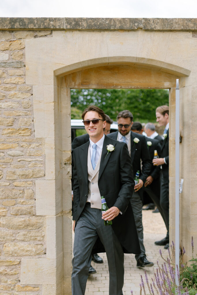 A smiling man in formal wedding attire and sunglasses walks through a stone archway holding a beer, followed by other men dressed in suits, some also holding drinks.