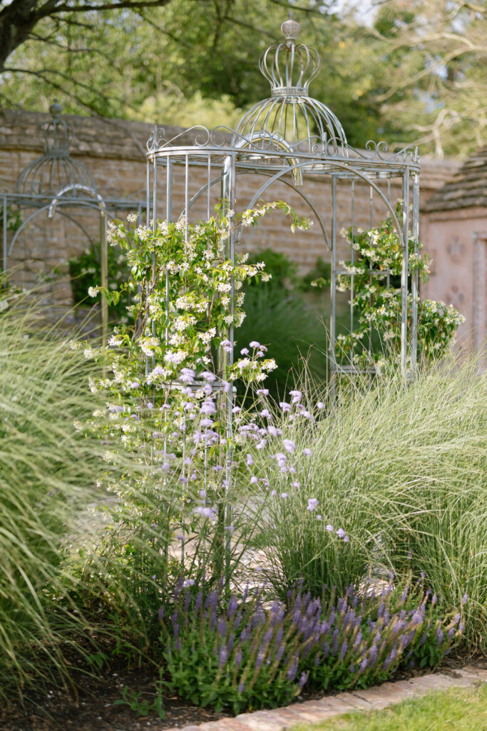 A metal gazebo covered with climbing white flowers is surrounded by tall grasses, purple and white flowers, and set in a lush garden with a stone wall in the background.