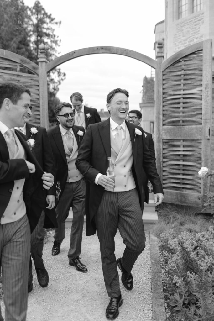 A group of groomsmen in formal suits laugh and walk outside through a wooden gate. One man in front, holding a bottle, smiles broadly. The scene appears candid and joyful.