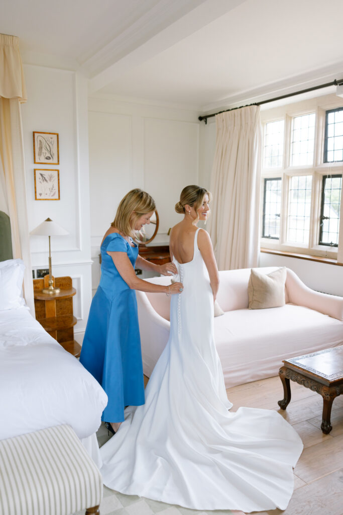 A woman in a blue dress helps a bride in a white gown button up her dress in a bright, elegant room with a sofa, bed, and large window.