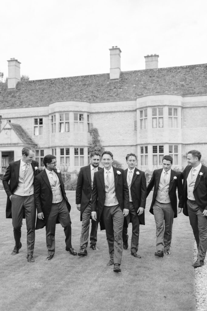 Seven men in formal suits walk together on grass in front of a large stone building with tall windows. The photo is in black and white, and the men appear to be smiling and enjoying themselves.