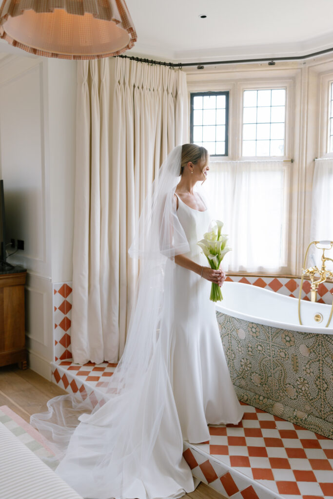 A bride in a white gown and veil stands by a bathtub, holding a bouquet of calla lilies, looking out a window with sheer curtains in a bright, elegant bathroom.