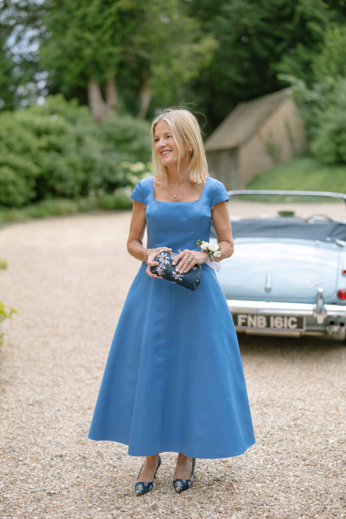 A woman in a blue dress stands outdoors on a gravel path, holding a clutch, with a vintage blue convertible car and greenery in the background.