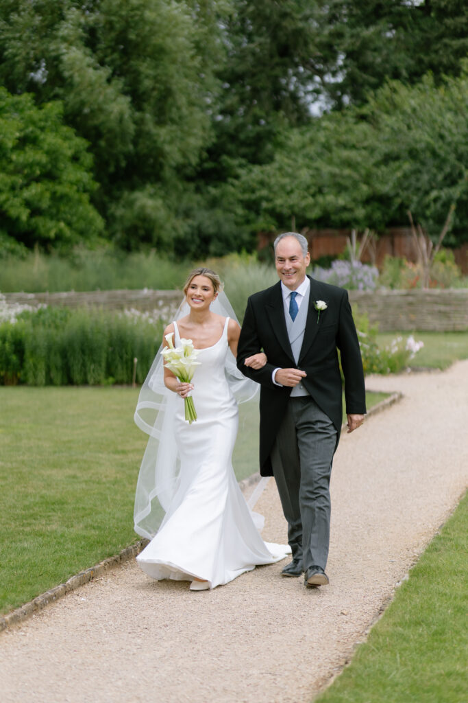 A smiling bride in a white dress walks down a garden path arm-in-arm with a man in a suit, surrounded by greenery.