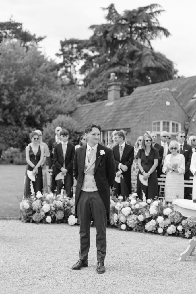 A groom stands outdoors in front of guests at a wedding ceremony, surrounded by flowers, with trees and a house in the background.