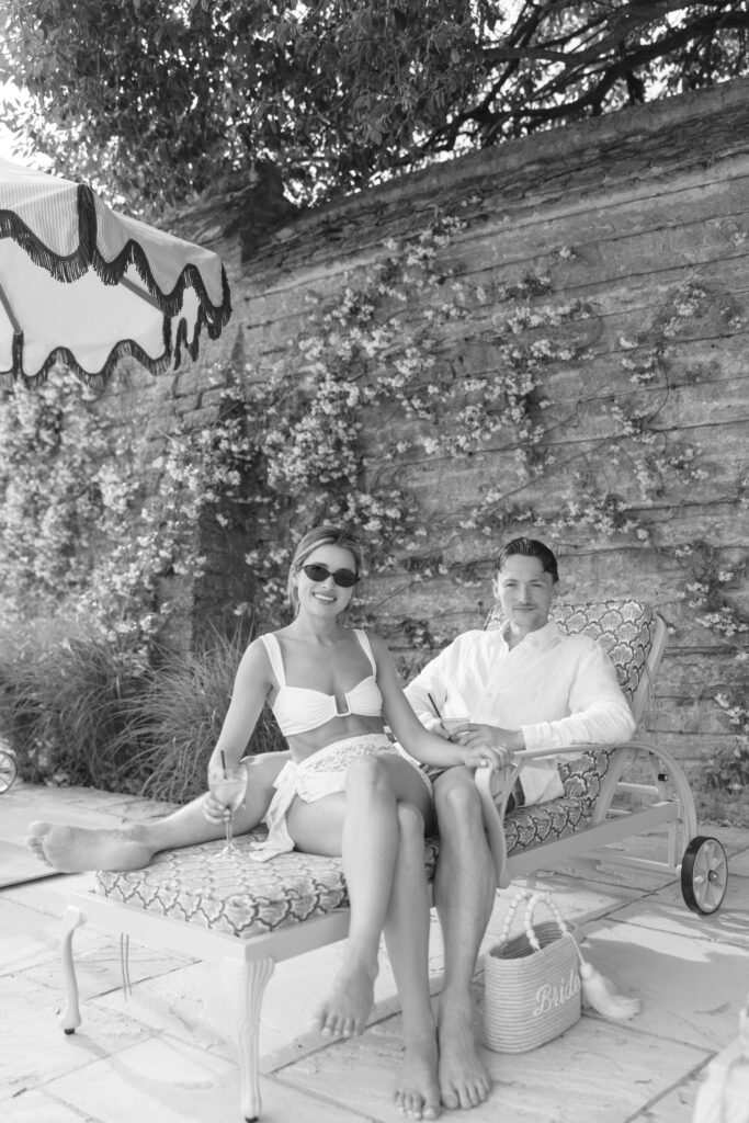 A couple sits together on a lounge chair by a pool. The woman wears sunglasses and a swimsuit, and the man is in a light shirt. Flowers climb the stone wall behind them. A basket labeled Bride sits nearby.